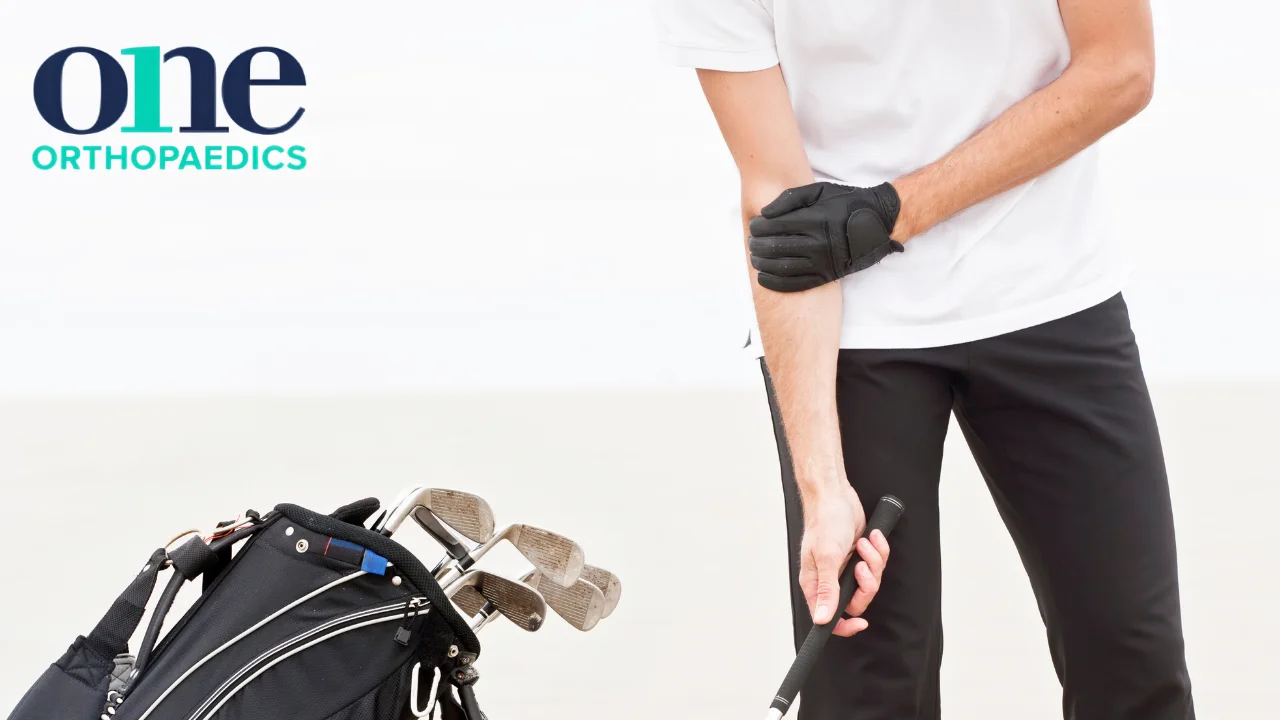 A person holding their elbow region in what appears to be discomfort due to golfer's elbow with a bag of golf clubs beside them. OneOrthopaedics logo is superimposed onto this image.