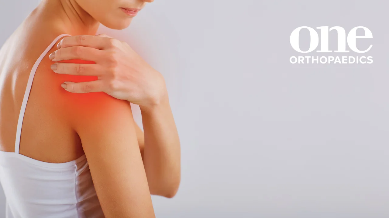 Picture of person holding their right shoulder in pain, with a One Orthopaedics logo placed on the top right-hand side of the image.